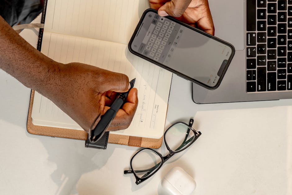 Overhead view of a man writing notes while using a smartphone and laptop, enhancing productivity.