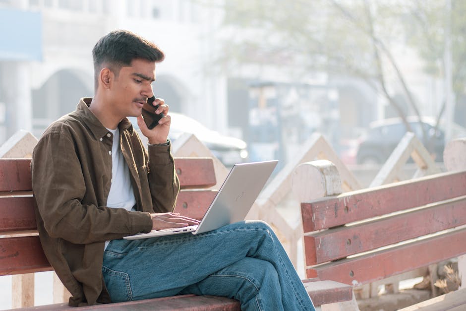 A young man multitasks with a laptop and phone on an outdoor bench, showcasing remote work.