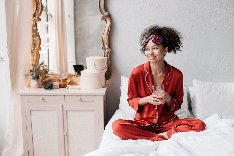 Smiling woman in cozy pajamas with curly hair enjoys a glass of water in a chic bedroom setting.