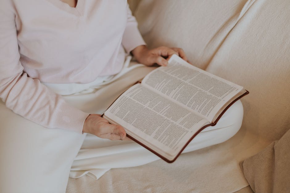 A woman sitting indoors on a couch reading an open book, wearing soft colored clothing.