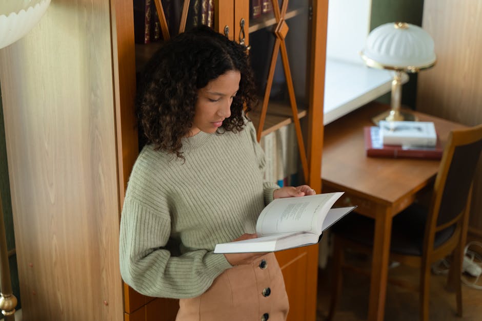 A young woman with curly hair absorbed in reading a book indoors by a bookshelf.