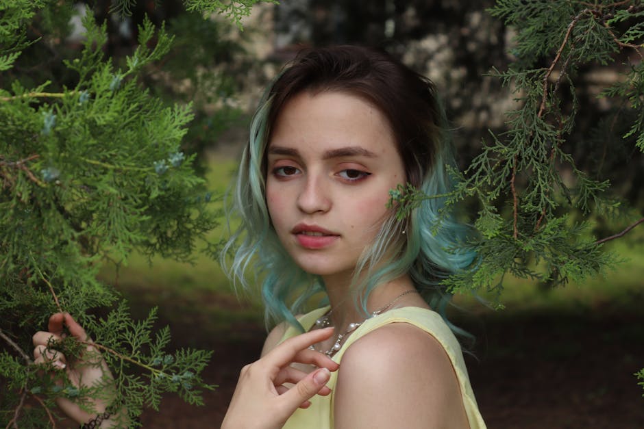 A young woman with green hair poses among green foliage outdoors.