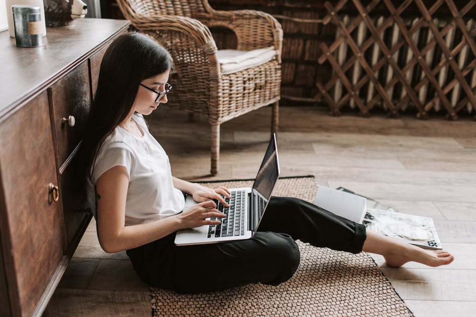 Side view of happy young woman with long dark hair in eyeglasses typing on keyboard of computer while sitting on floor in modern living room